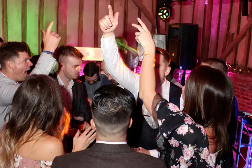 Wedding party crowd dancing with hands raised in barn venue under pink and red uplighting