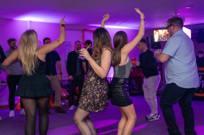 Group of women dancing with hands raised under pink purple uplighting at evening hen party